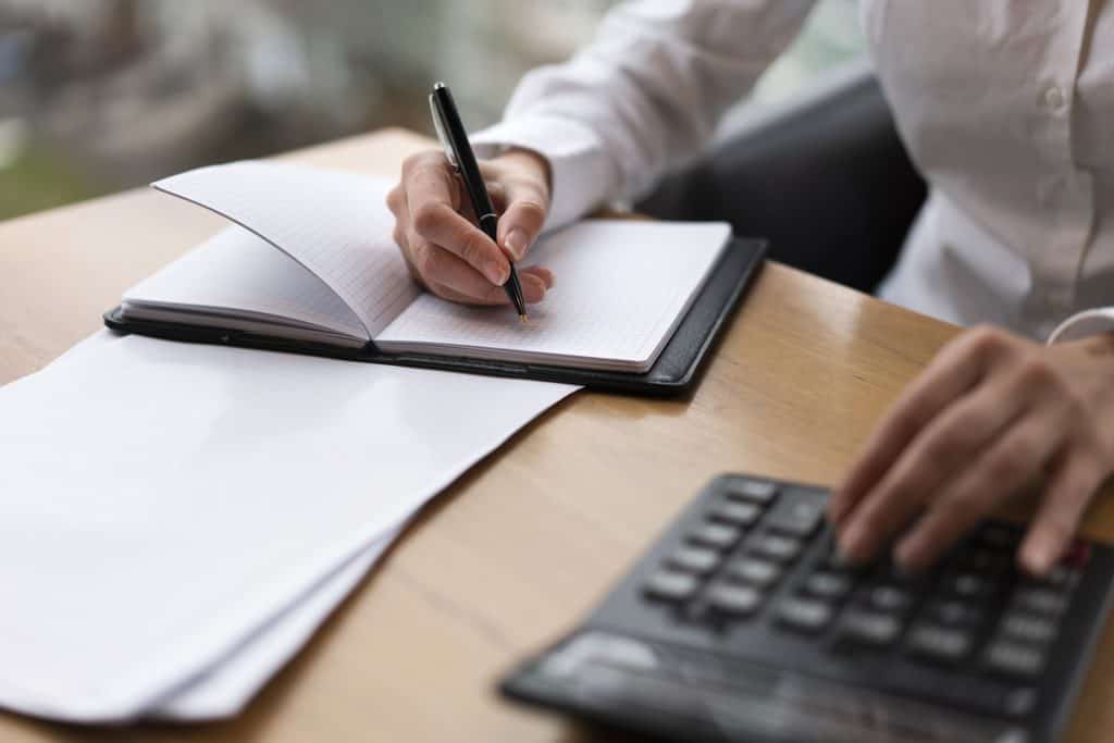 Close-up of a person in a white shirt writing in an open grid notebook with a black pen while the other hand presses the keys of a black calculator, with loose papers on a wooden desk.