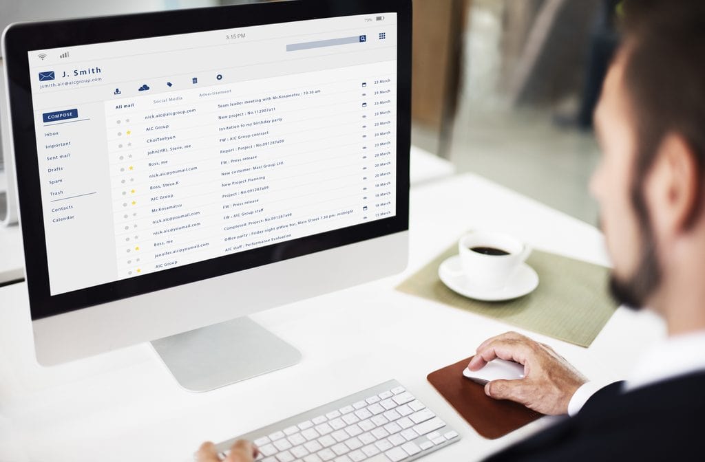 Man in a dark suit using a mouse and keyboard at a white desk, looking at a desktop monitor that shows an email inbox with a compose button, folders, and a list of messages, beside a cup of coffee on a saucer.