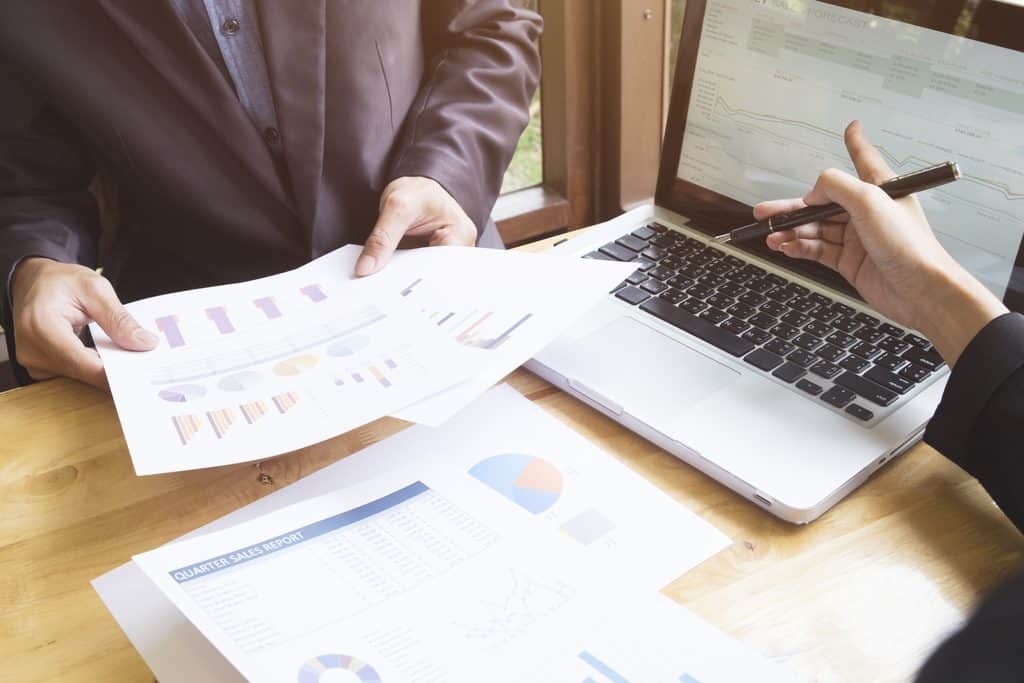 Two people seated at a wooden table, one holding printed charts and graphs while the other points a pen at a laptop screen showing a sales forecast, with a quarter sales report and pie chart on the table.