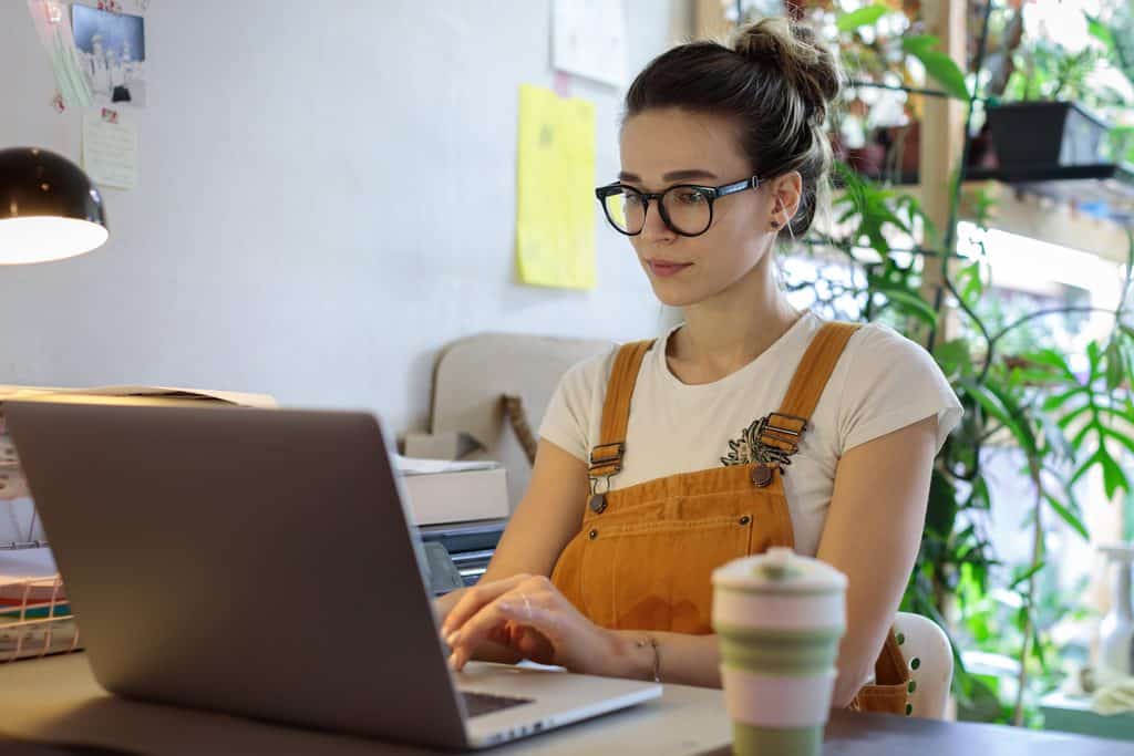 Woman wearing glasses and orange overalls typing on a laptop at a desk with a lamp, a reusable coffee cup, and plants around her, researching the best portfolio websites freelancers can build.