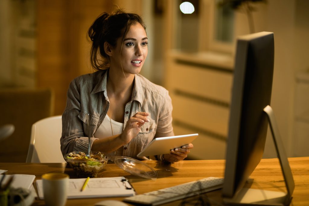 Woman with her hair in a bun smiling and pointing toward a desktop monitor while holding a tablet at a wooden desk with a bowl of salad, a coffee mug, a clipboard with sketches, and a yellow pencil, reviewing her work for Upwork Profile Optimization.