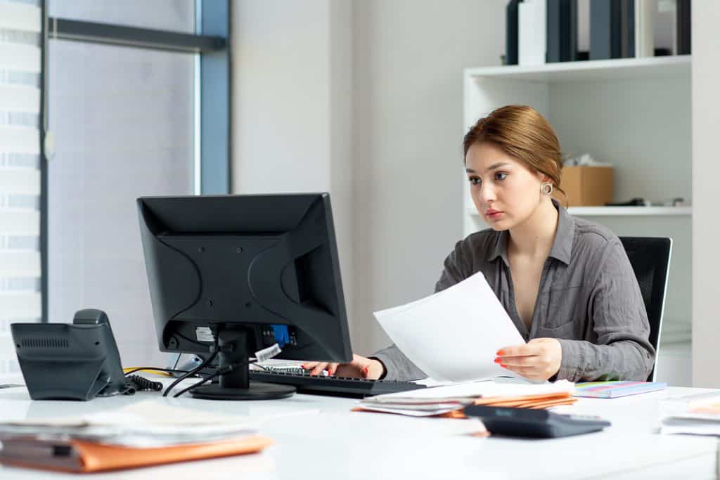 Woman with her hair tied back, wearing a gray button-up shirt, holding a white paper in one hand and typing on a keyboard with the other at a desktop computer, with a desk phone, stacked folders, and a calculator on a white desk while checking payment terms for Should Freelancers Charge Deposits.