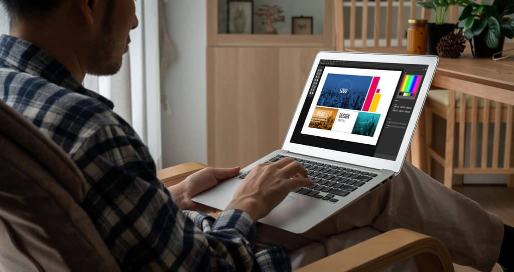 Man in a blue plaid shirt sitting in a wooden armchair and typing on a silver laptop that shows design software with a logo layout, an ideas panel, a color palette, and a layers panel.