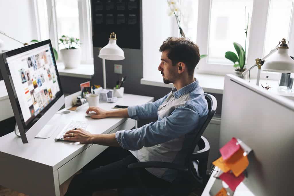 Man in a blue denim shirt sitting at a white desk and looking at a large desktop monitor showing a grid of photos, with a lamp, plant, coffee cup, phone, and sticky notes nearby, illustrating a Freelance Web Design Guide workspace.