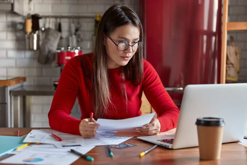 Woman with long brown hair, round glasses, and a red sweater holding a pen and reading a paper document at a kitchen table with an open laptop, scattered papers, markers, and a takeaway coffee cup