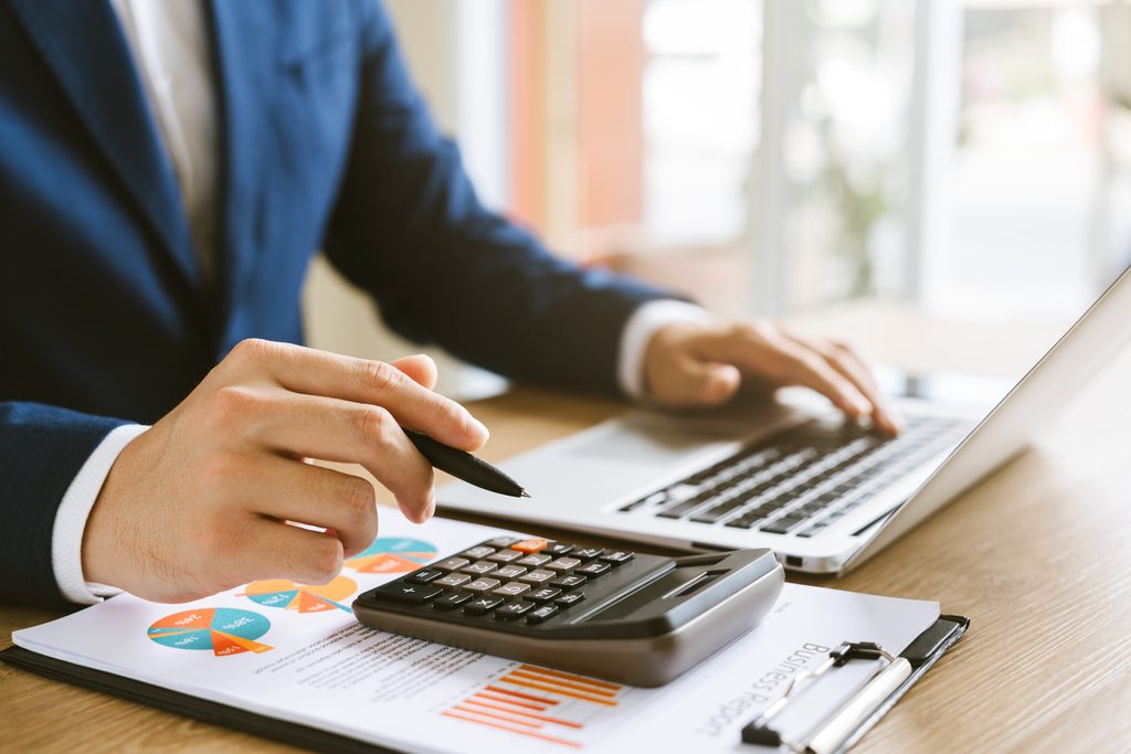 Close-up of a person in a navy suit holding a black pen over a handheld calculator that rests on a business report with colorful pie charts and bar charts, while the other hand rests on a silver laptop keyboard, with a clipboard on a wooden desk.