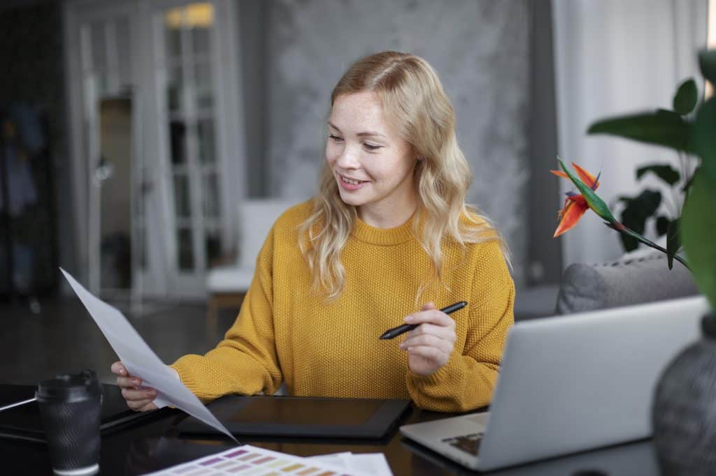 Smiling blonde woman in a mustard knit sweater holding a printed sheet and a stylus at a desk with a drawing tablet, an open laptop, a takeaway coffee cup, and color swatches in front of her.