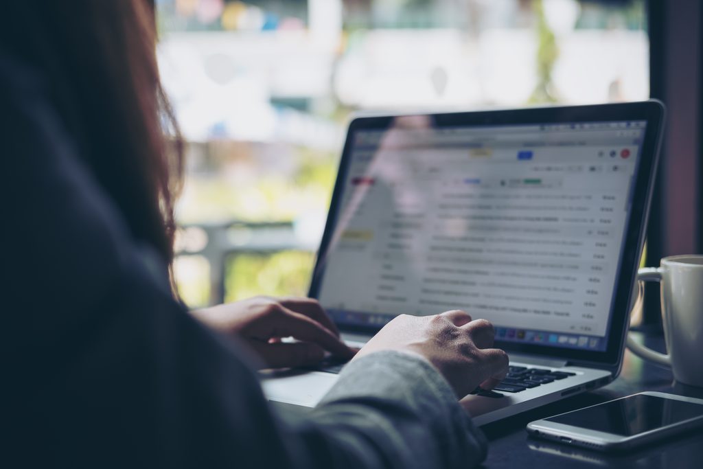Over-the-shoulder view of a person with long dark hair typing on a laptop that shows an email inbox, with a white coffee mug and a smartphone on a dark table beside a window, sending an agreement to a client as part of How to Create Freelancer Contracts.