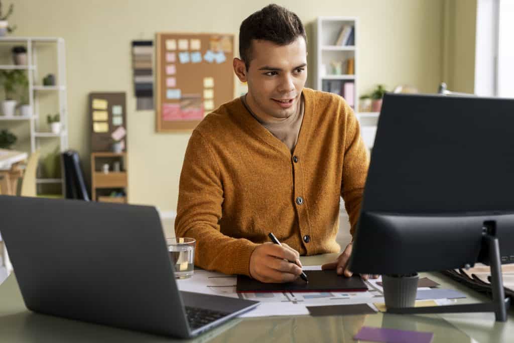 Man in an orange cardigan holding a stylus over a drawing tablet while looking at a computer monitor, with a laptop and a glass of water on the desk, showing a freelancer at work in a Freelancing Versus Remote Job comparison.