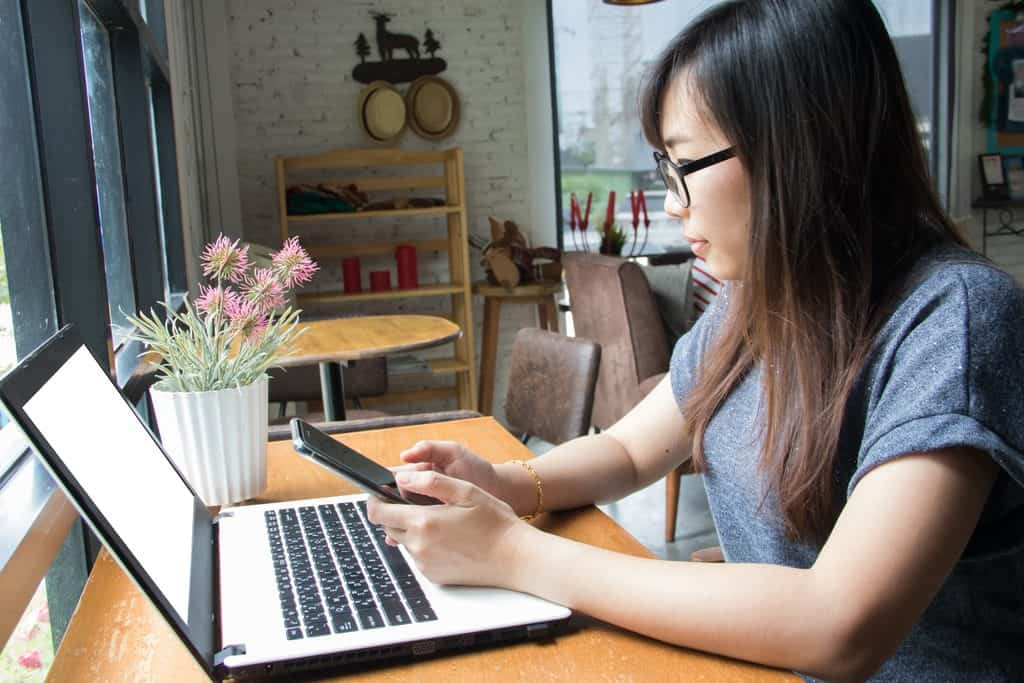 Woman with long hair and black-framed glasses holding a smartphone beside an open laptop with a blank white screen on a wooden table by a café window, with a pot of pink flowers nearby.