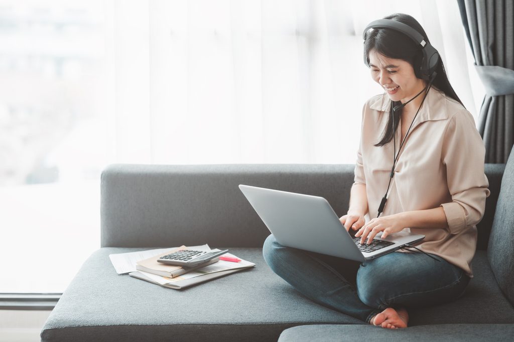 Woman wearing a headset and smiling while typing on a laptop cross-legged on a sofa, with a calculator and notebook beside her.