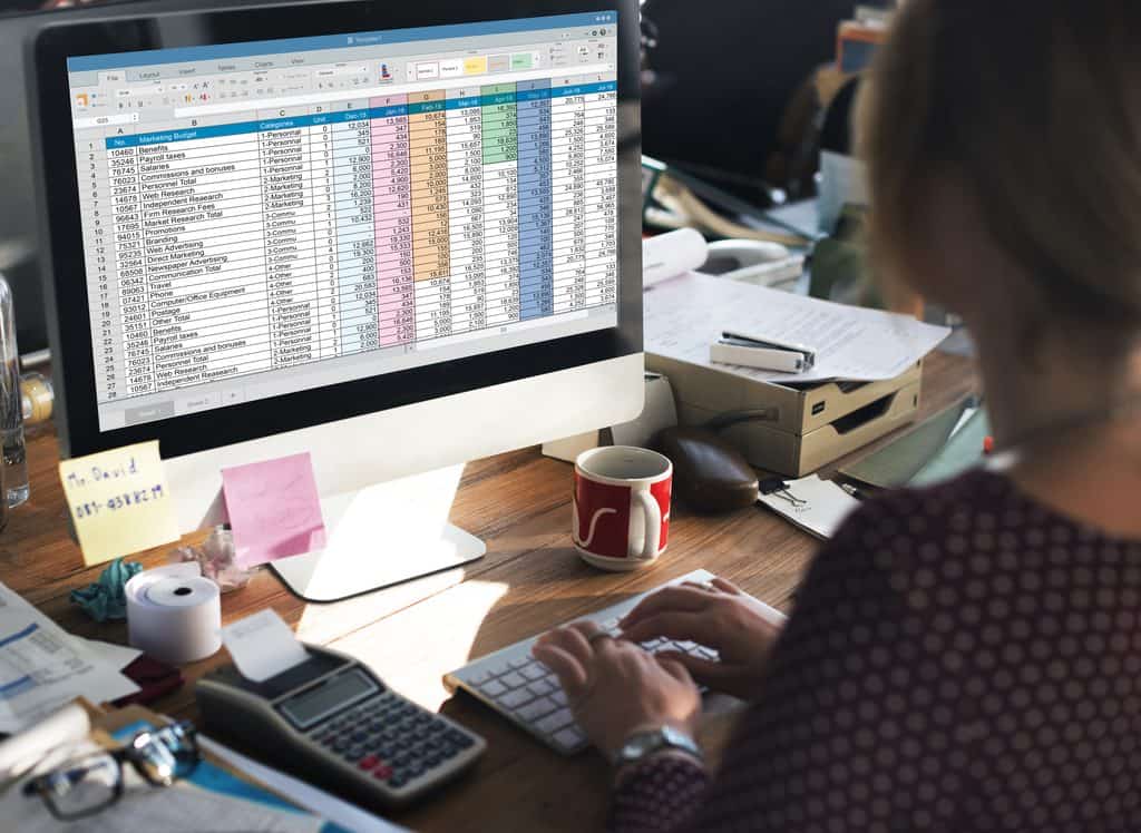 Woman typing at a desktop monitor displaying a color-coded Marketing Budget spreadsheet with monthly columns, beside a printing calculator, sticky notes, a red coffee mug, and stacked papers