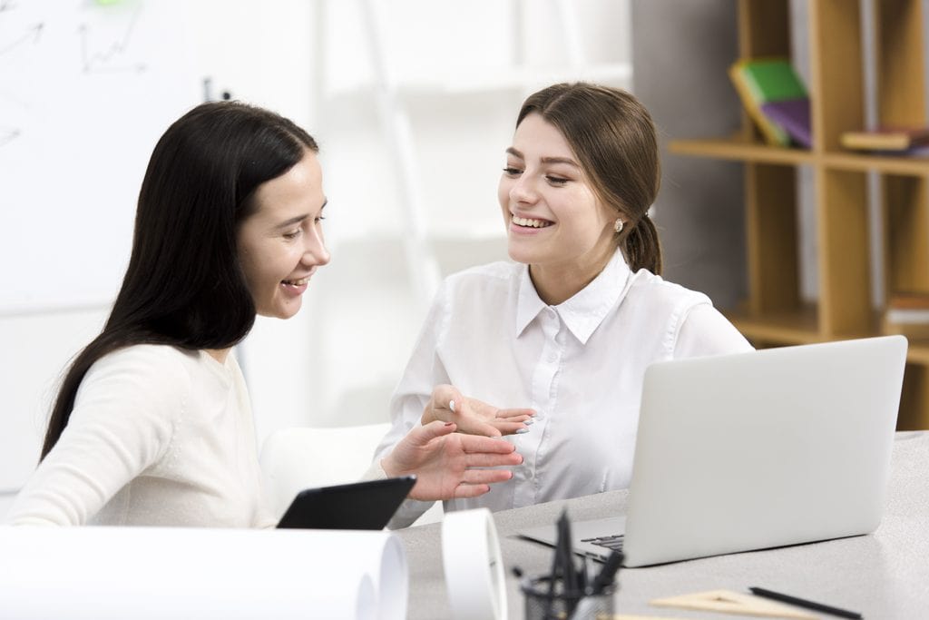 Two smiling women in light tops sitting at a desk with an open laptop, a black tablet, and rolled papers, talking and gesturing while discussing the Freelancing Versus Remote Job question.