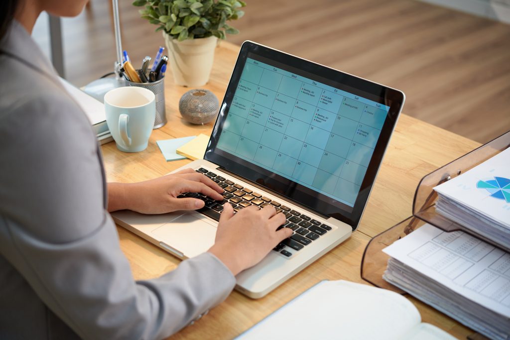 Woman in a blazer typing on a laptop displaying a monthly calendar with color-coded meetings and tasks, with a stack of papers beside her.