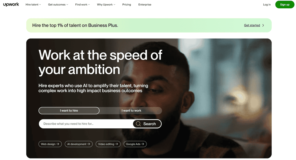 Upwork homepage with the headline Work at the speed of your ambition, an I want to hire and I want to work toggle, a search box, and shortcut buttons for web design, AI development, video editing, and Google Ads over a photo of a man’s face.