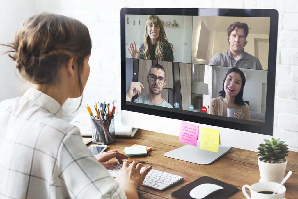 Woman with her hair in a bun working at a desktop computer during a four-person video call, with stationery, sticky notes, and a coffee cup on the desk.