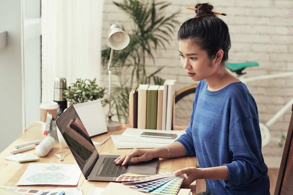 Woman with dark hair in a bun held by a pencil, wearing a blue sweater, working on a laptop with one hand while holding a fan of color swatches, with printed patterns, markers, books, and a lamp on a wooden desk