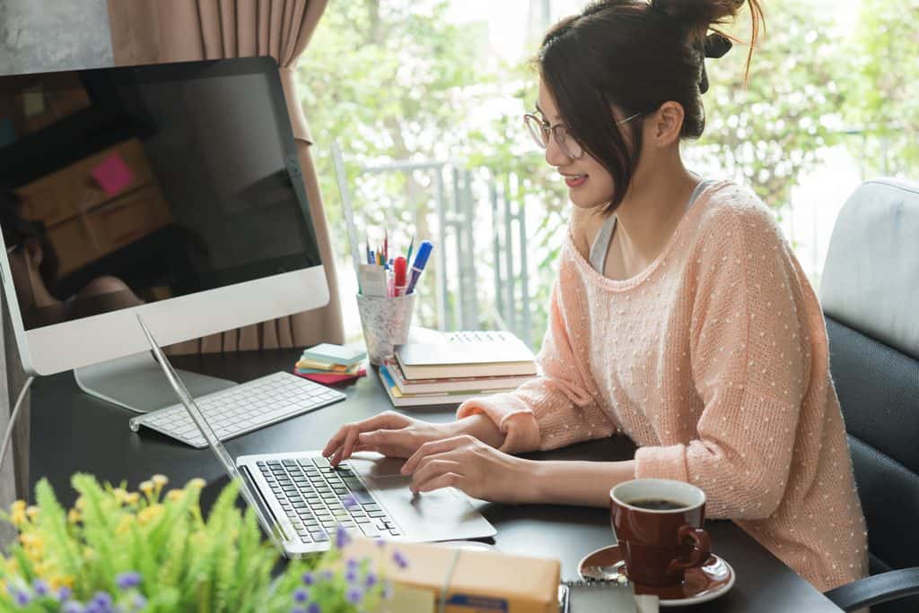 Woman with glasses and her hair in a bun, wearing a peach polka-dot sweater, smiling and typing on a laptop at a dark desk with a desktop monitor, stacked books, sticky notes, a coffee cup, and a small plant in a Freelance Web Design Guide home office.