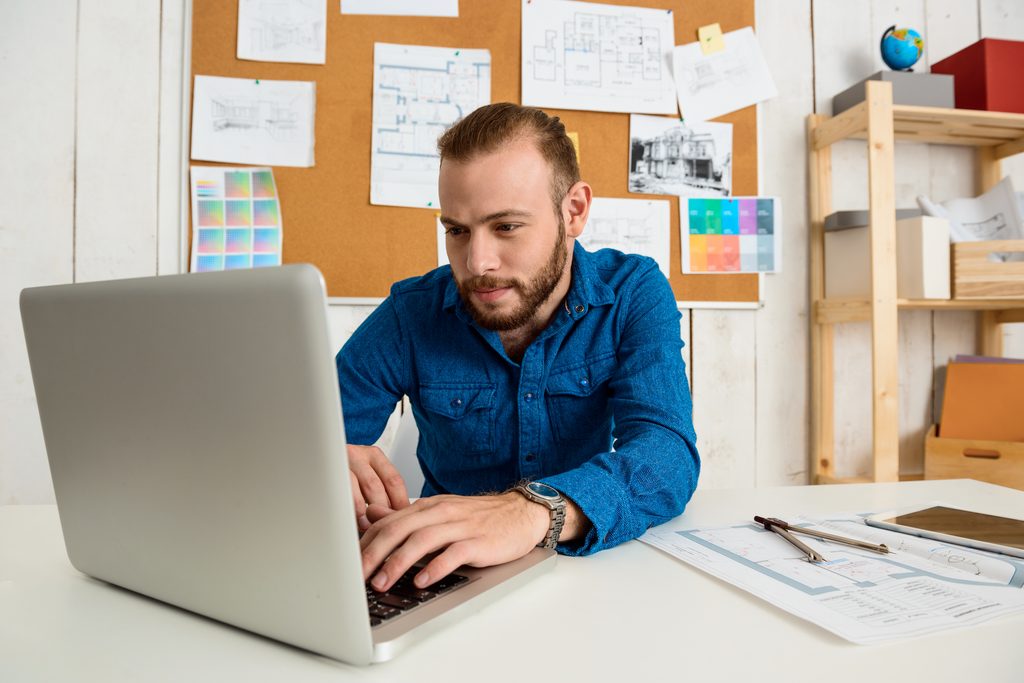 Bearded man in a blue denim shirt typing on a silver laptop at a white desk, with a corkboard of floor plans, sketches, and color swatches behind him and a drafting compass, drawings, and a tablet on the desk.