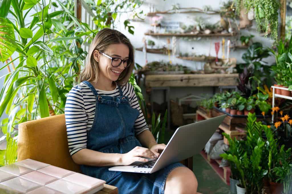 Smiling woman with glasses, a striped shirt, and a denim overall dress working on a laptop in an orange armchair, surrounded by green plants and wooden shelves.