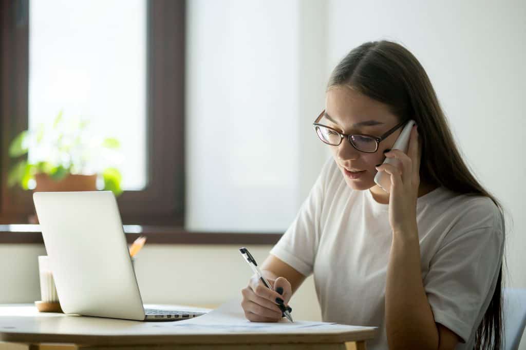 Woman with long brown hair and glasses, wearing a white t-shirt, talking on a phone while writing notes with a pen on paper at a table beside an open laptop, near a window with a potted plant, as part of building a Service Based Side Hustle.
