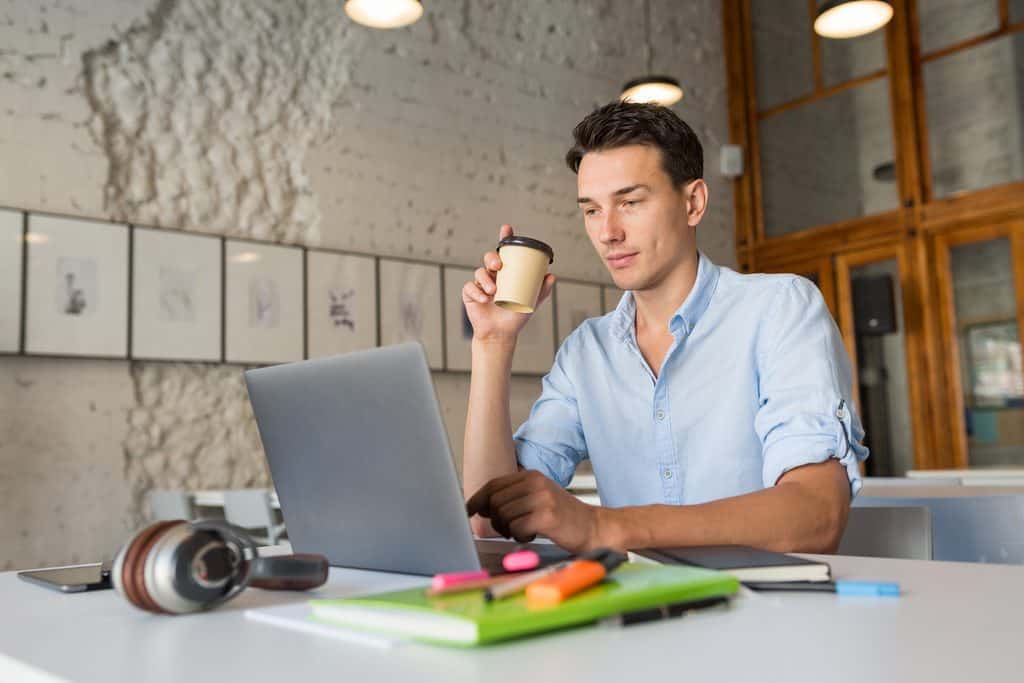 Young man in a light blue button-up shirt holding a takeaway coffee cup and looking at a silver laptop at a white table, with headphones, a green notebook, and highlighter markers in front of him in a loft-style workspace with framed art on the wall.
