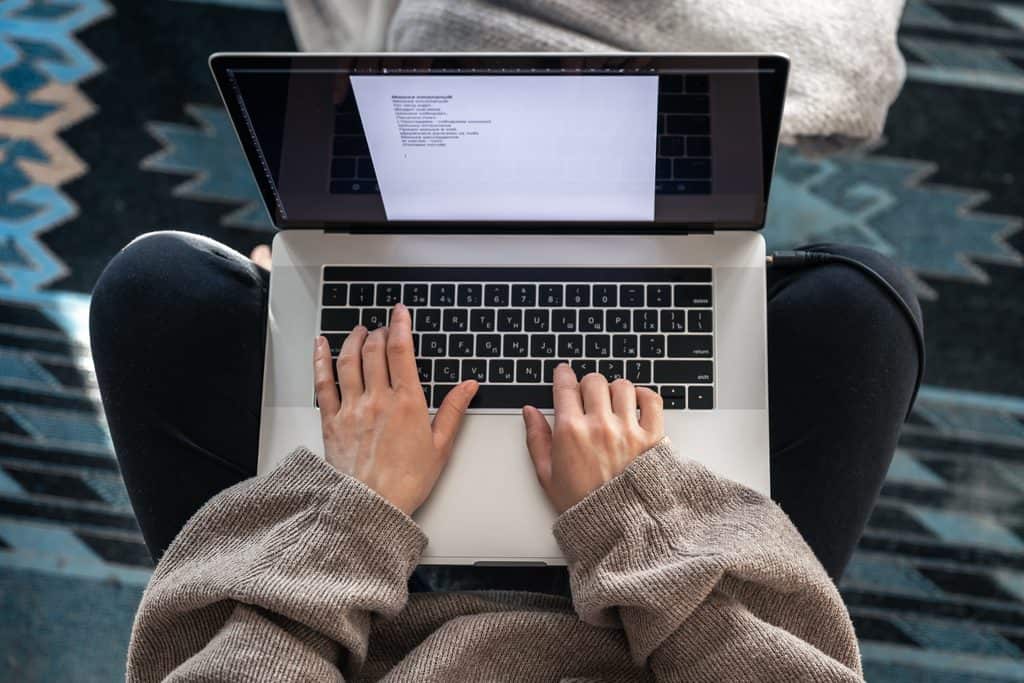 A woman in a beige knit sweater and dark pants sitting cross-legged on a patterned blue rug, typing on a laptop, top view